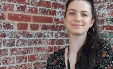 Image of Harriet smiling at the camera sat in front of an exposed brick wall background