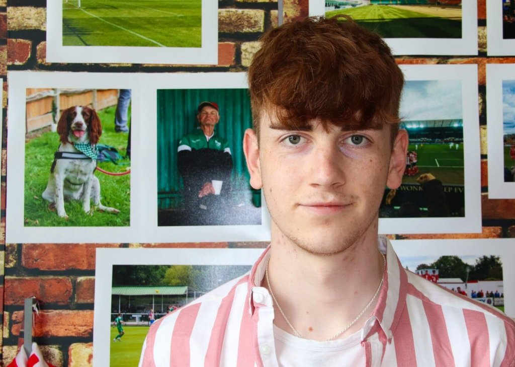 Image of Harry head and shoulders view wearing a pink and white stripe shirt stood in front of a display of his photography work