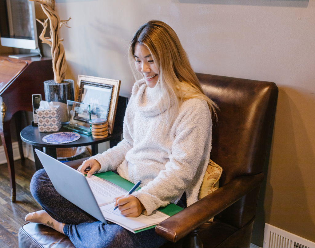 Smiling young woman sat cross legged on chair working with laptop on lap