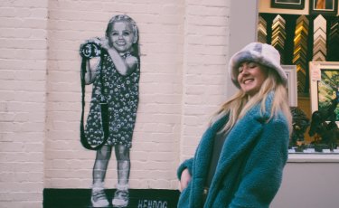 a girl with blonde hair wearing a hat with turquoise jacket is standing in front of a wall art of an little girl in black and white