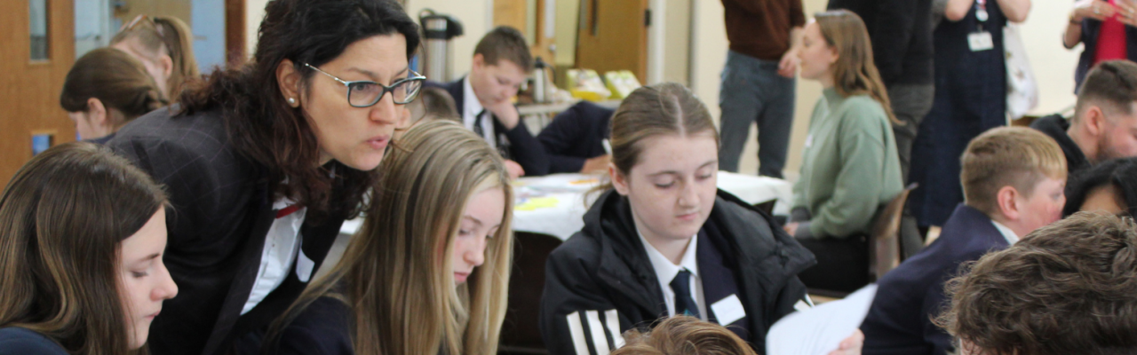 Angmering School EXPO 22/23. A woman leaning over a table full of young people to explain something. image